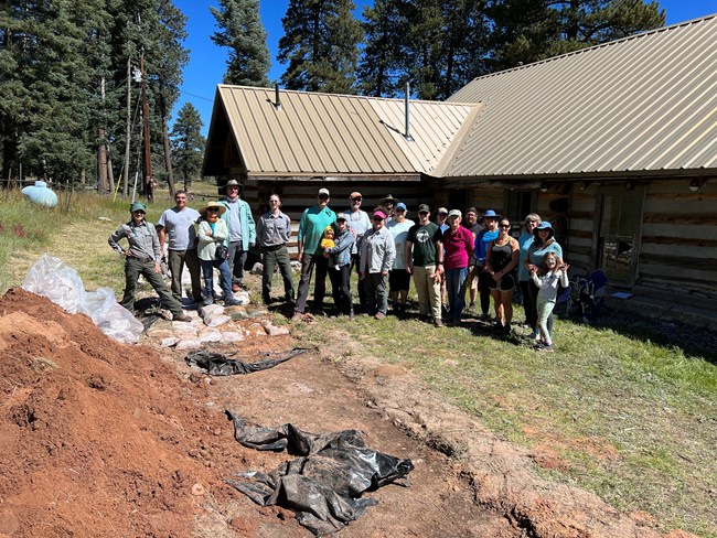 A large group of volunteers and park employees stands in a group and smiles at us from behind a historic cabin.