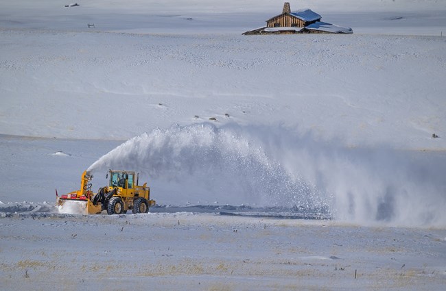 A yellow snow plow blows a horizontal plume of snow off of a roadway in a vast, snowy landscape.