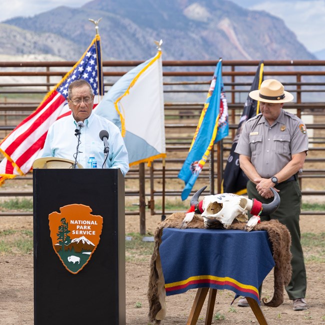 a person bowing their head in prayer at a podium in front of Tribal and U.S. flags at a bison facility