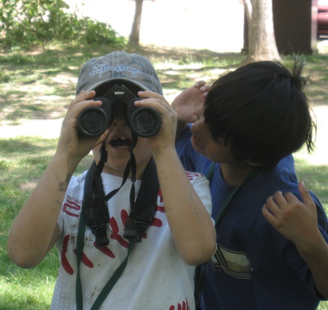 Montezuma Well Junior Ranger Camp Birding A boy looks through binoculars with gaping mouth as a second boy tries to look over his shoulder.