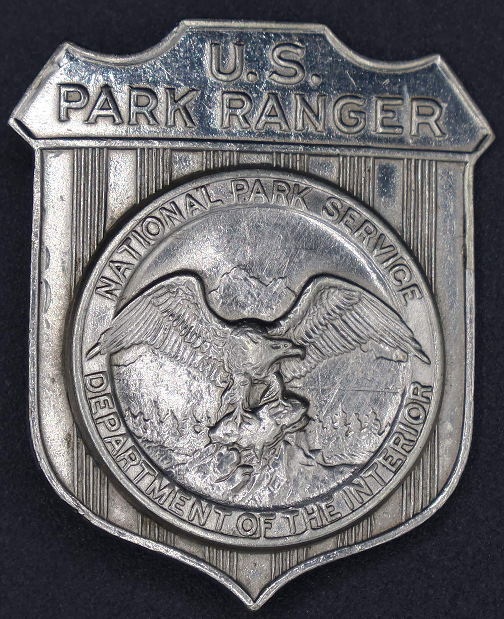 Silver shield-shaped badge marked U.S. Park Ranger. The raised round seal in the middle has an eagle looking to its left.