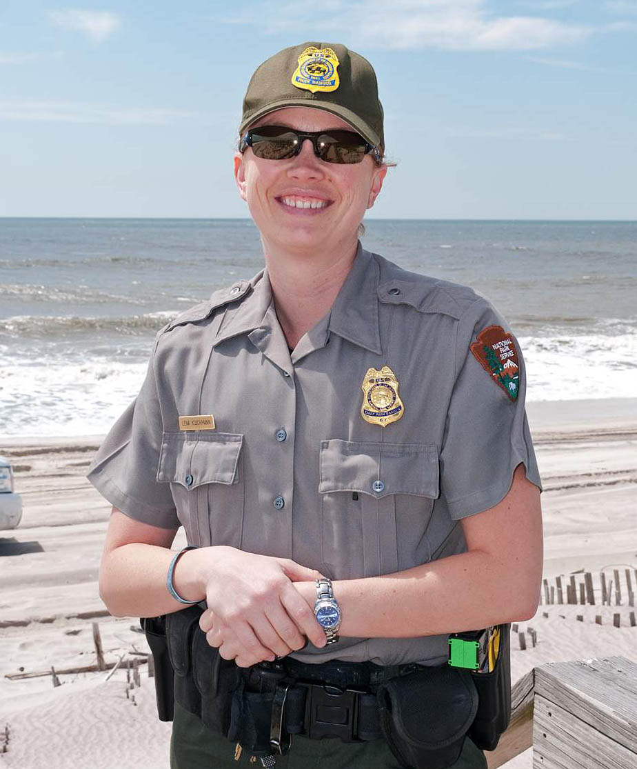 Lena Koschmann stands on a beach wearing NPS uniform and ball cap. She has a shield-shaped badge with an eagle perched on top pinned to her shirt.