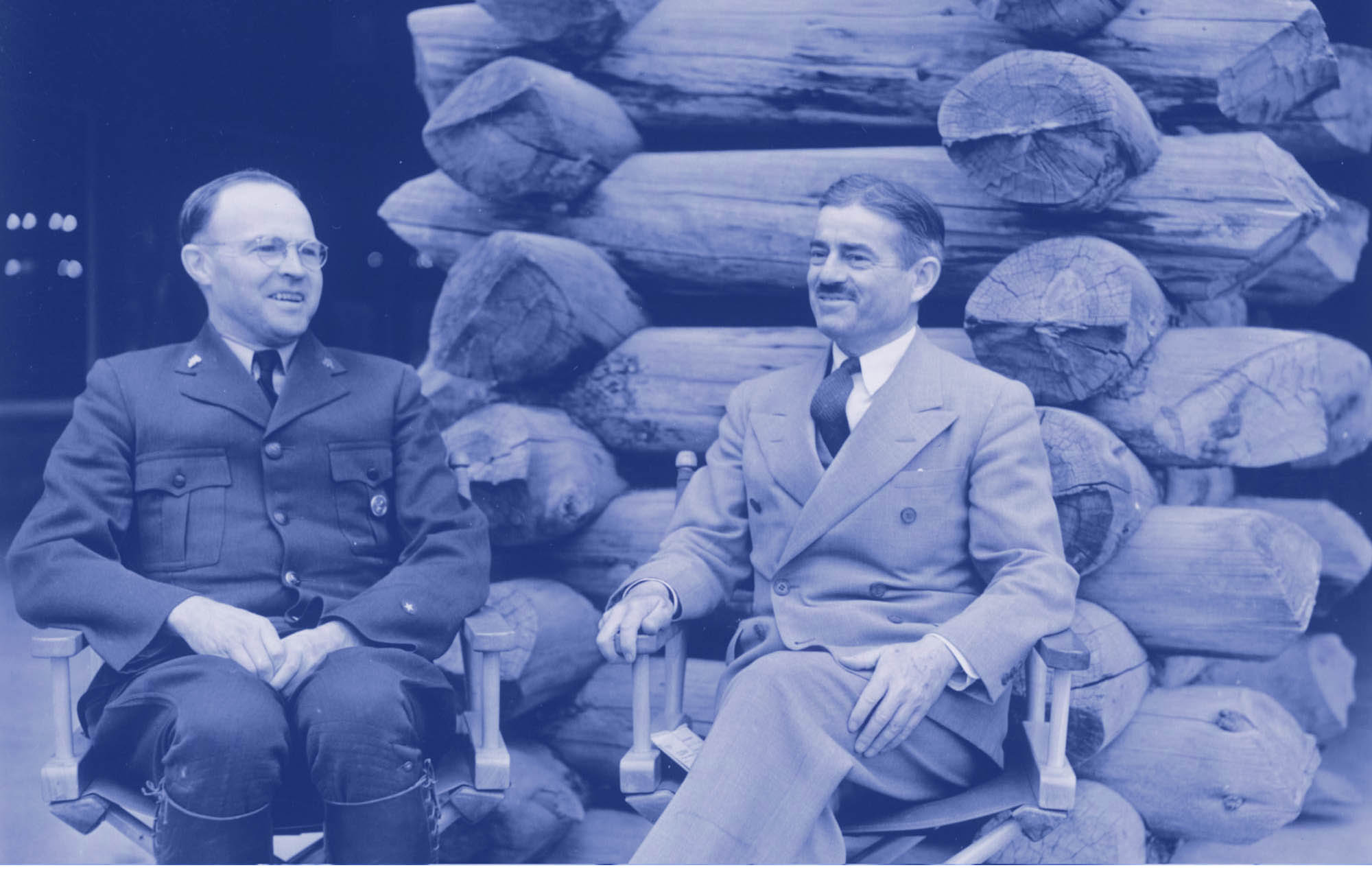 Edmund Rogers and Newton Drury sit in front of a stack of logs. Rogers wears the NPS uniform with a round badge. Drury wears a suit.