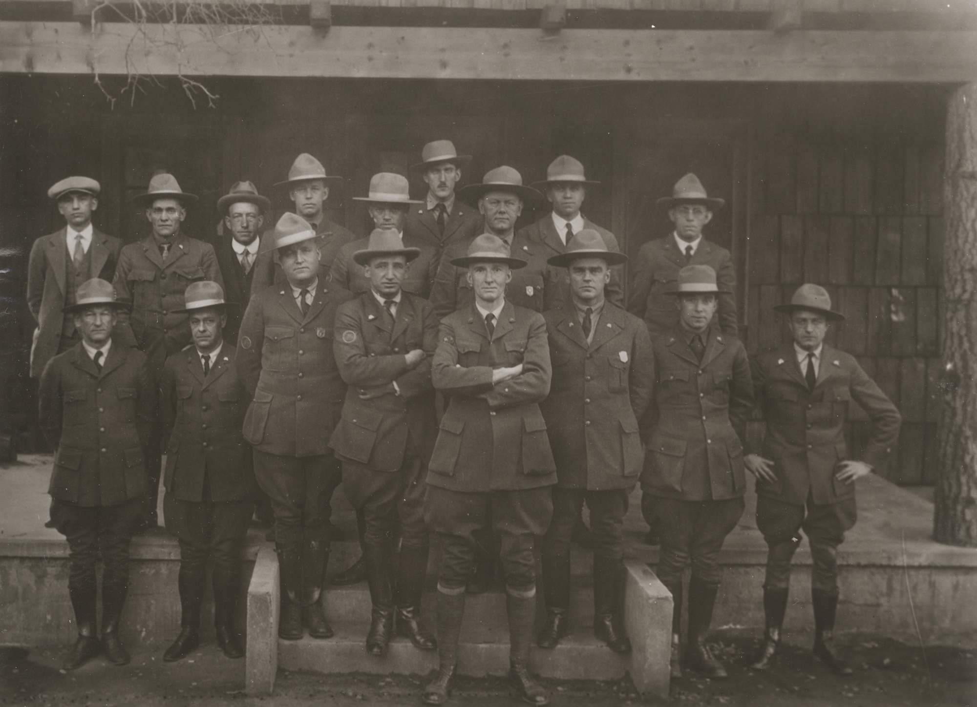 Seventeen men pose in in front of a building, 15 in NPS uniforms with broad brim hats. Shield-shaped badges are worn only by three men.