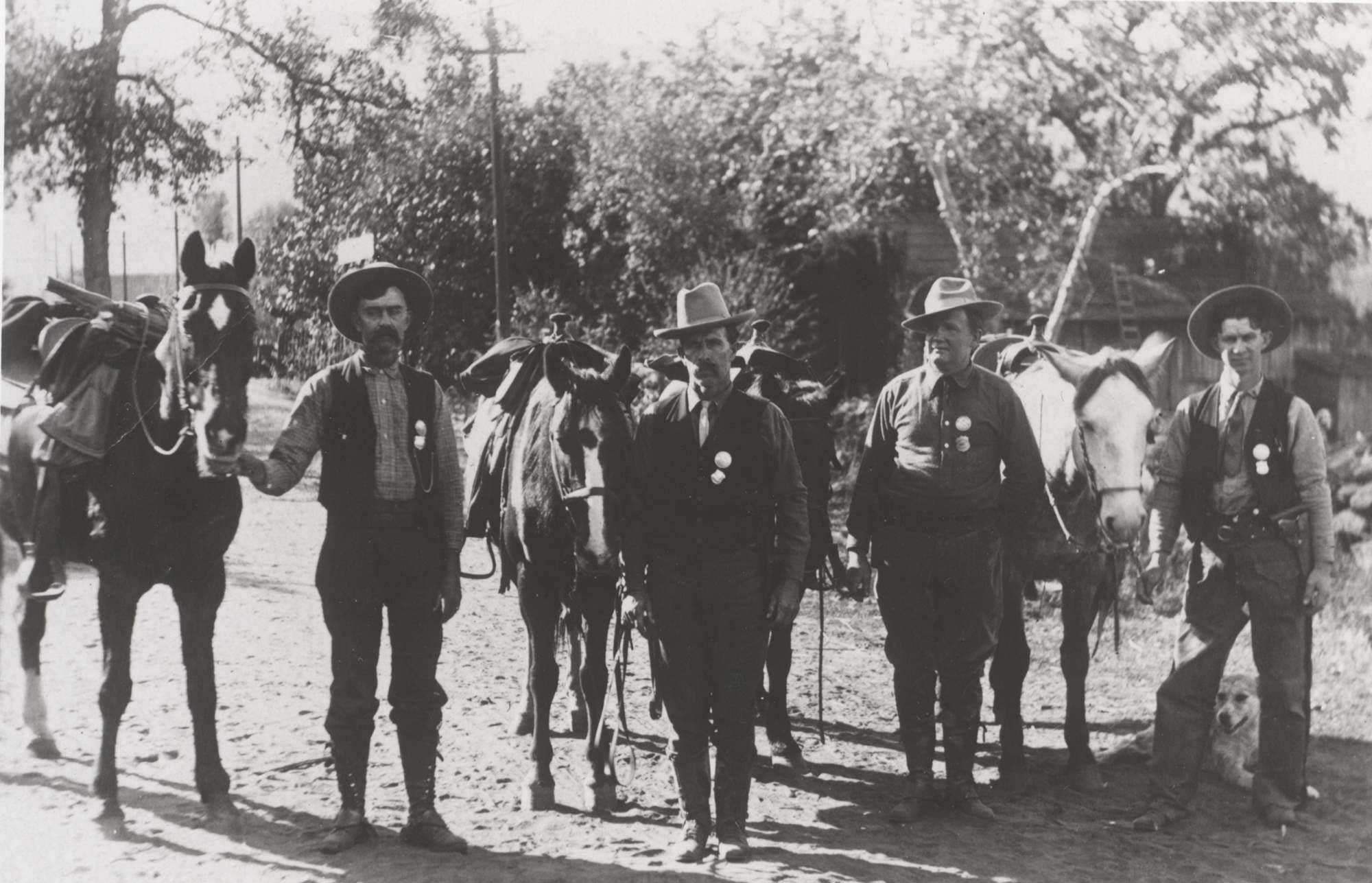 A man stands by another on horseback. Both wear breeches, boots, shirts, and flat brim hats. One has a round badge on his shirt.