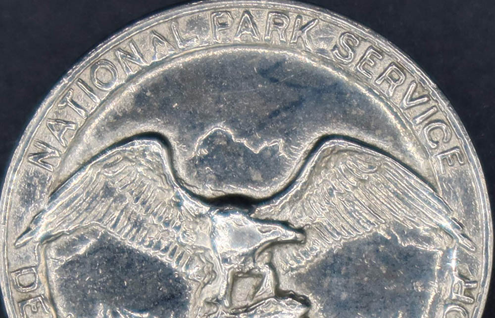 Round silver badge with National Park Service Department of the Interior around the rim and an eagle looking to its left in the middle.