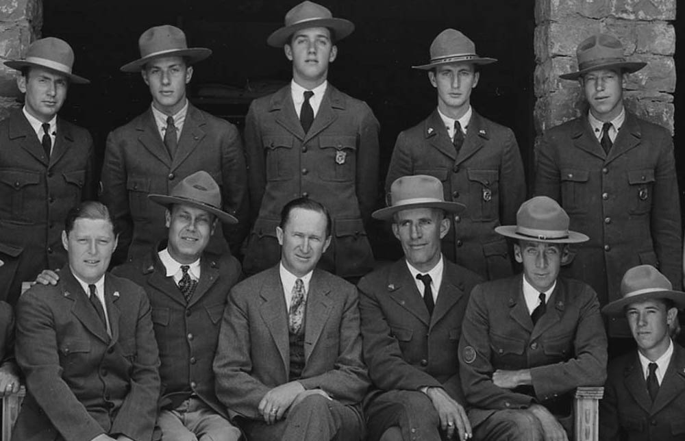 Seventeen men and a woman pose with Director Albright.  Shield shaped badges are on the jacket pockets of five uniformed rangers. 