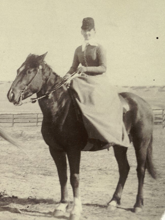 Kate Graham sits side-saddle on a dark colored horse. She wears a long dress buttoned to her throat. She wears a scarf around her throat and a dark colored riding hat.