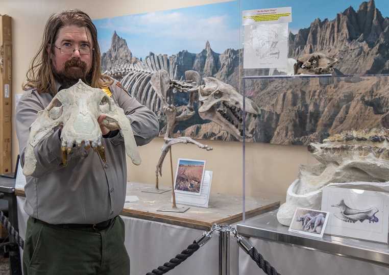 A ranger in uniform with shoulder length brown hair, mustache, goatee, and wire-rimmed glasses holds an animal skeleton's skull while standing indoors in front of a full model skeleton.