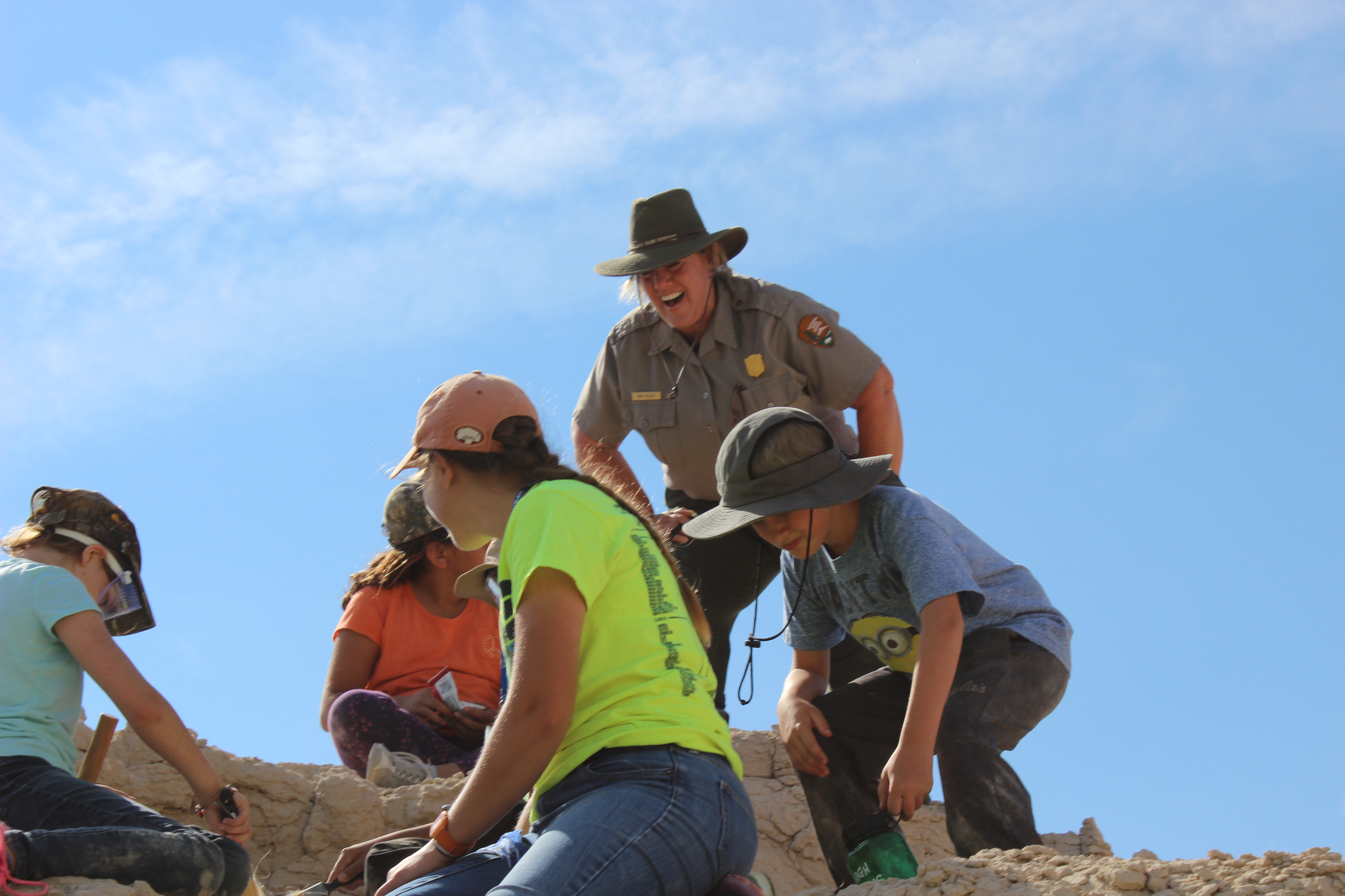 A blond ranger in uniform and hat observes 4 kids excavating fossils. .