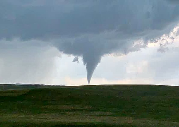 A dark gray tornado under gray clouds appears over green rolling hills.