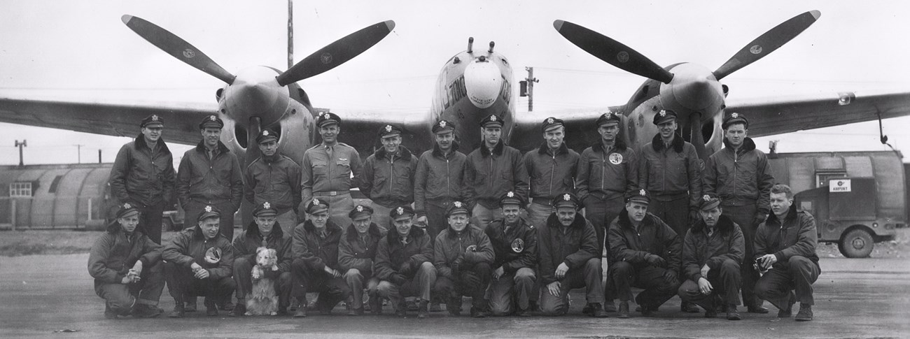 a historic scene of 23 uniformed men standing in front of an airplane with two large propellers. one soldier holds a dog.