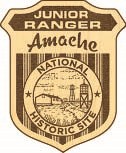 Amache NHS 3-Hi-Res-CROP wooden junior ranger badge reads "Junior Ranger" across the top. "Amache" is in script underneath. "National Historic Site" is in a circle around a scene depicting a barrack and guard tower.