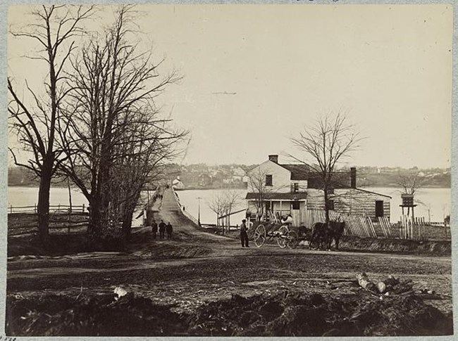 A black and white photo of a bridge across a river. A small wooden house is at the base of the bridge.