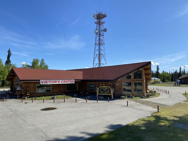 View of a large one-story angular log building. A large sign attached to the eaves reads "Visitor's Center". A large radio tower stands in the background against a clear blue sky