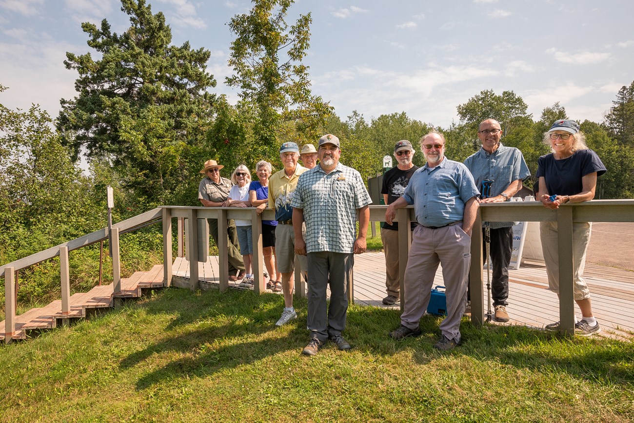 National Park Service Director with Friends of the Apostle Islands Photograph of group of people looking towards camera with a staircase to their right, trees in the background, and a blue sky.