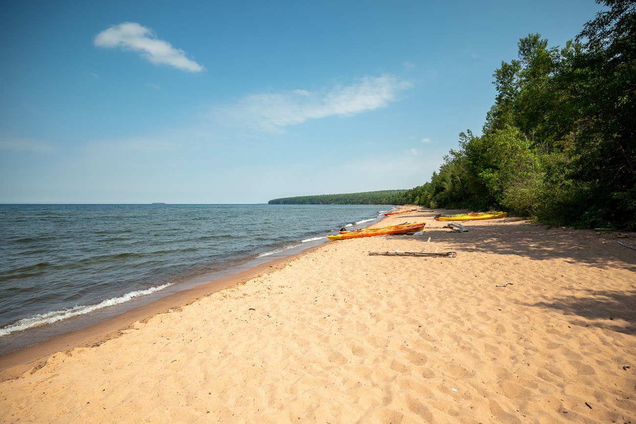 Photograph of a long sandy beach with Lake Superior on the left and trees on the right. Colorful orange and yellow kayaks line the sand.