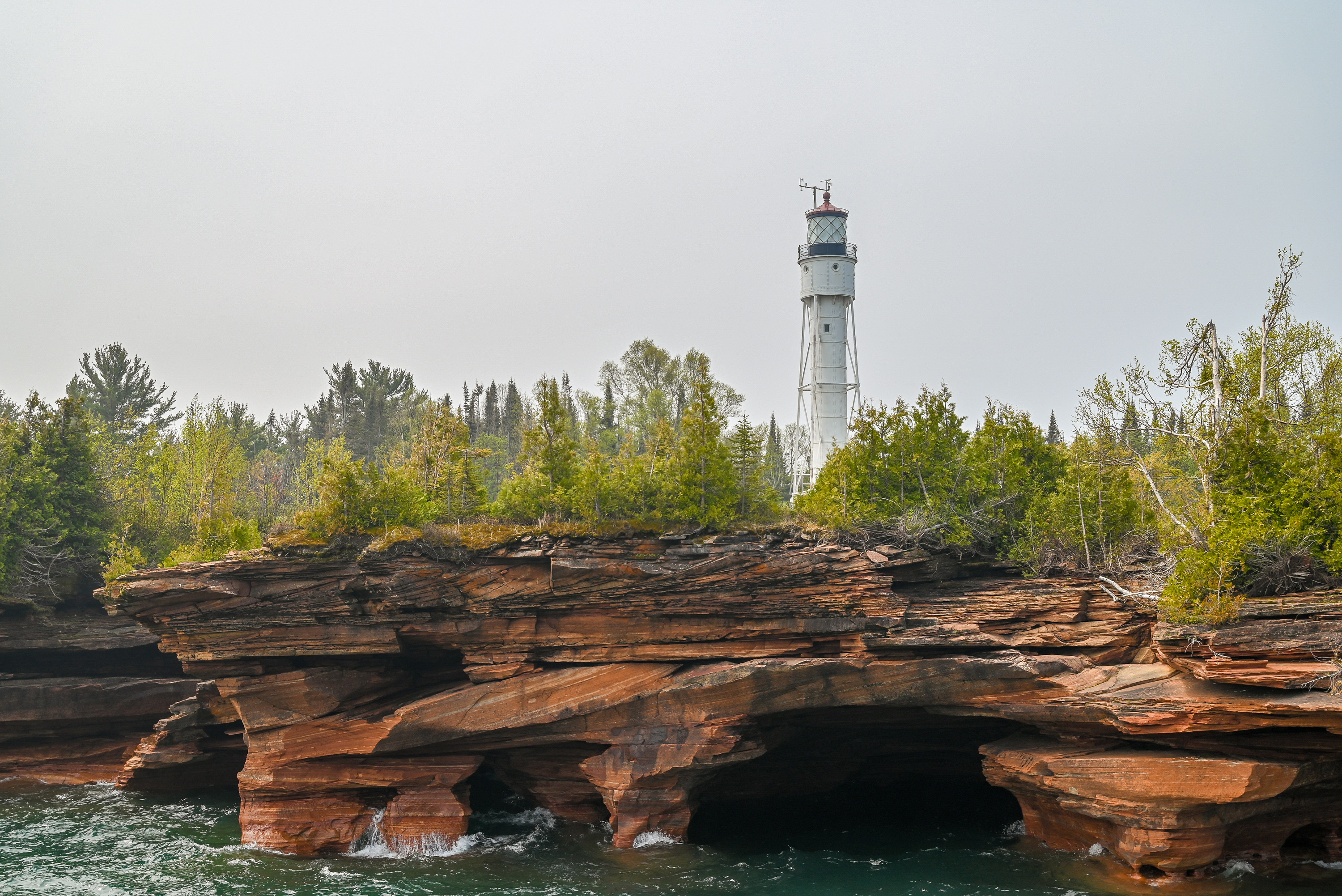 Photograph of lakeside sandstone cliffs with treelined shoreline and the top of a white lighthouse visible in the background.