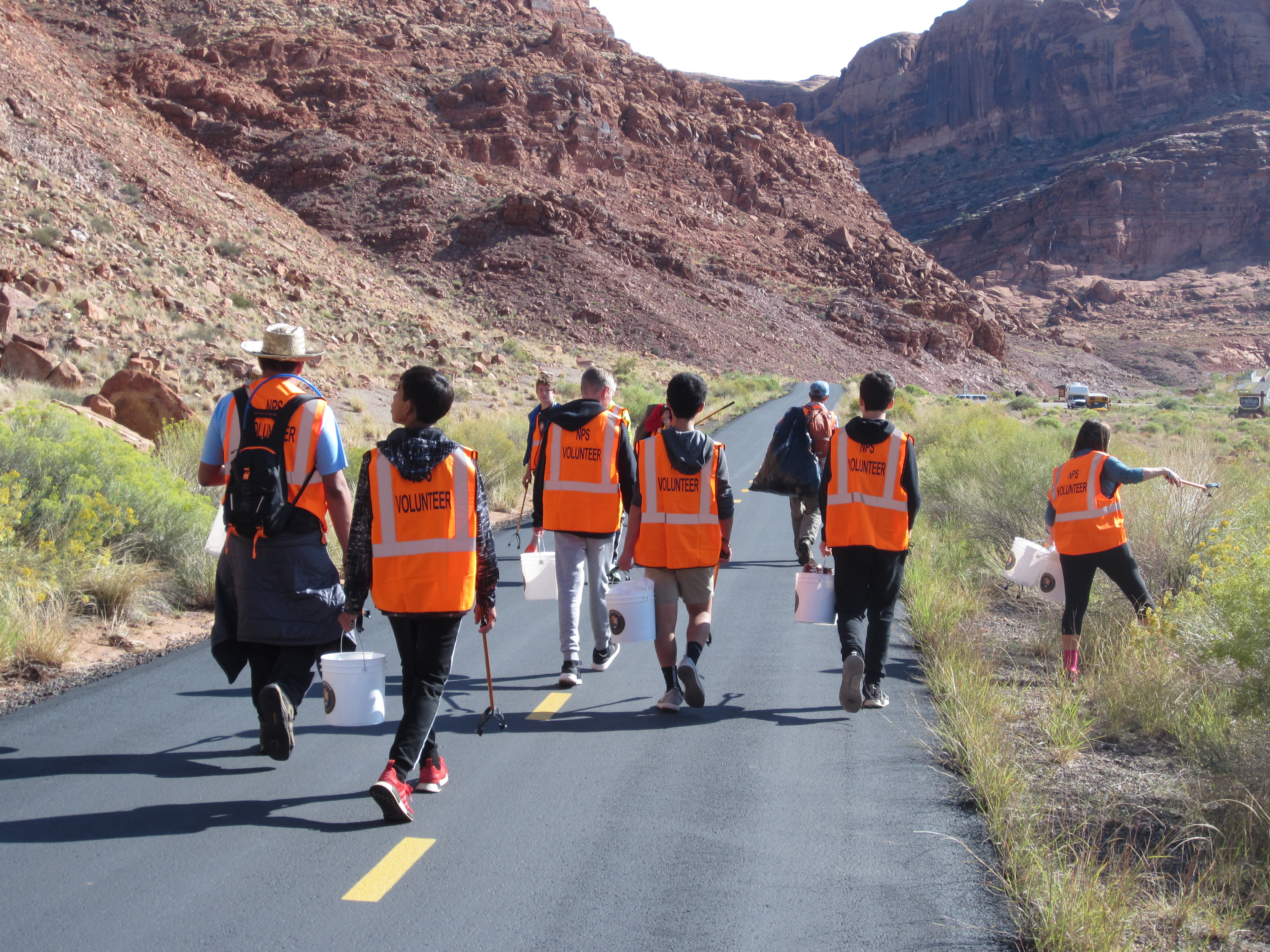 Volunteers in bright orange vests walk along a bike trail and pick up litter among the colorful cliffs of Arches National Park.