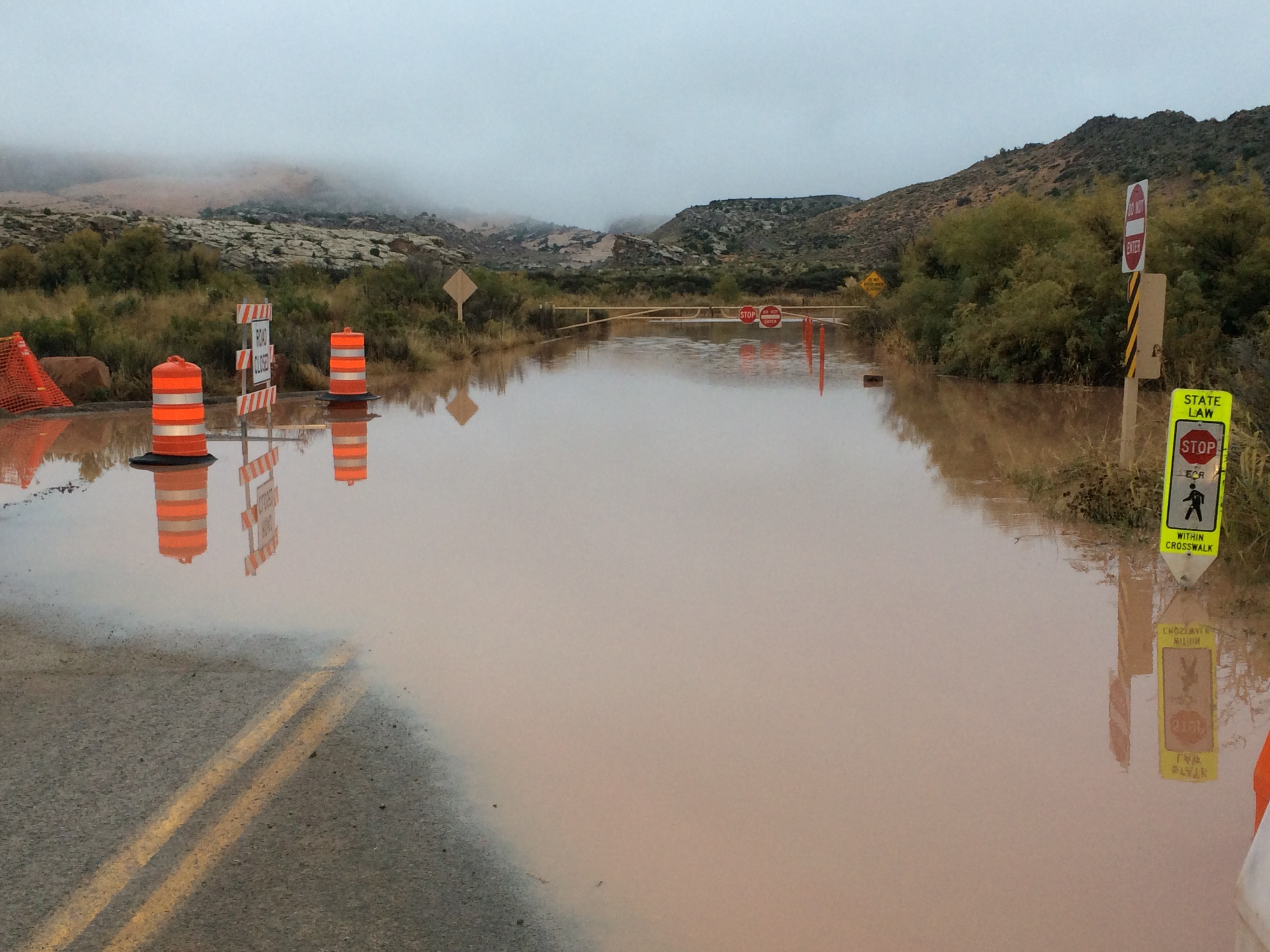 brown water covers a paved roadway for 50 feet in front of a barricade, low shrubs on roadsides