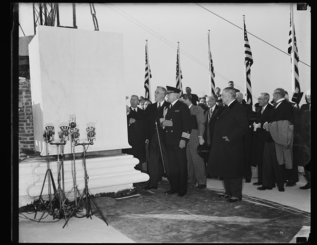 President Franklin Roosevelt with hand on marble slab of Jefferson Memorial cornerstone surrounded by men in suits and holding arm of man in military uniform. Four American flags on flagpoles behind crowd, five news microphones in front