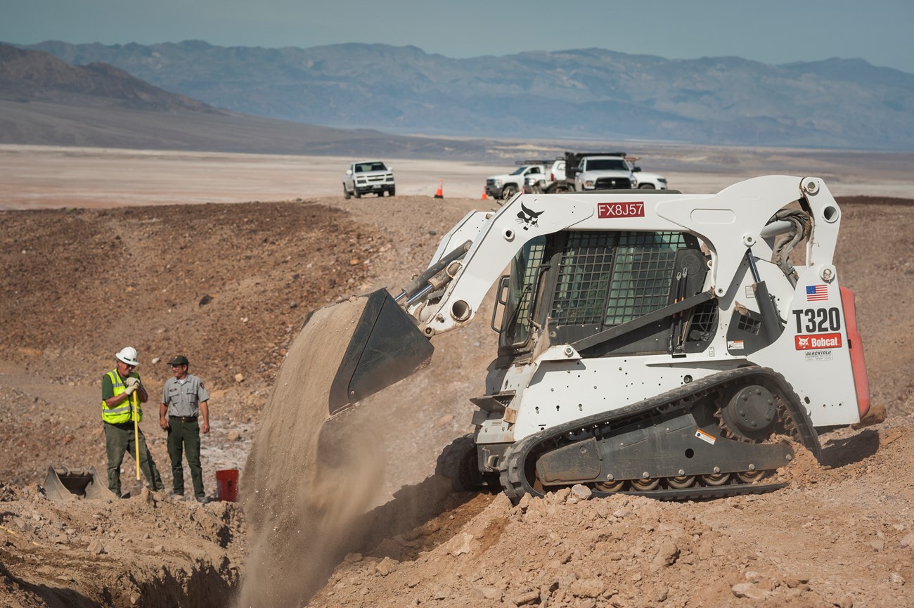 A white machine dumps dirt into a trench. Men in uniform and reflective vests stand to the left. The landscape is bare open ground with distant mountains.