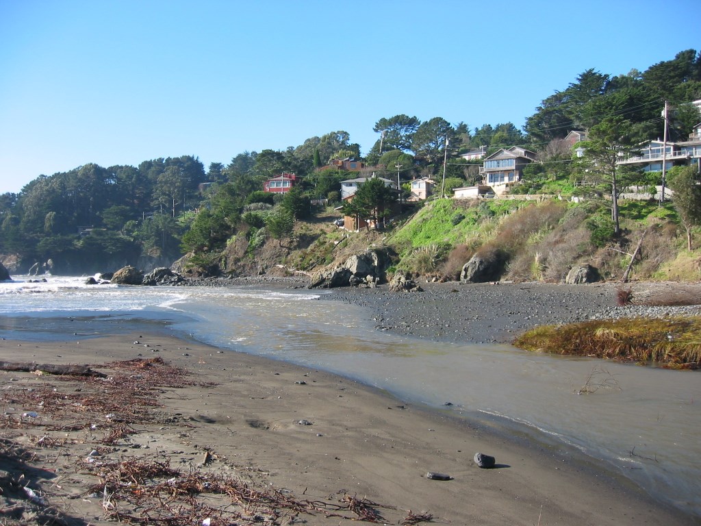 Creek crossing a beach and meeting the ocean just below a bluff topped with rows of homes.