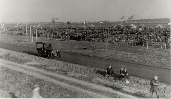 Black and white image of a grassy airfield with stands set up. A large amount of individuals are seated in the spectator stands. An old car sits in the left side of the image on a paved road. A few people appear in the lower right of the image.