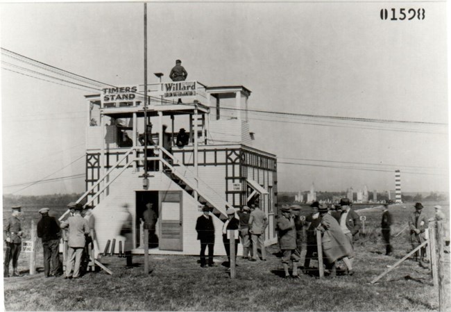 Black and white image of a timers stand set up on a grassy field. There are many individuals standing in the foreground of the image in front of the stand. There are wires and a pole near the stand.