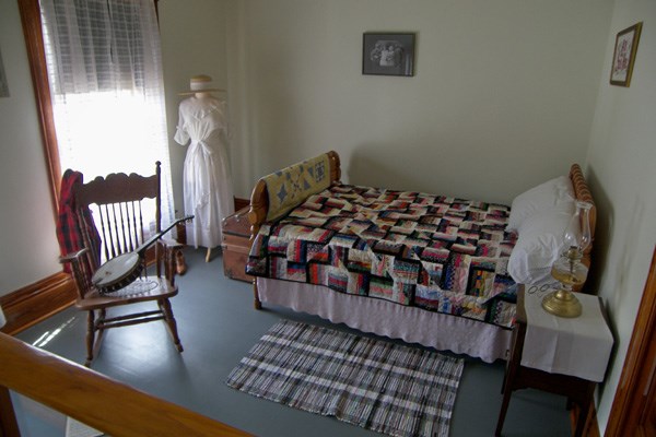 A full size bed covered with a colorful quilt, a rocking chair with a banjo on it, and a white dress hanging up in the corner.