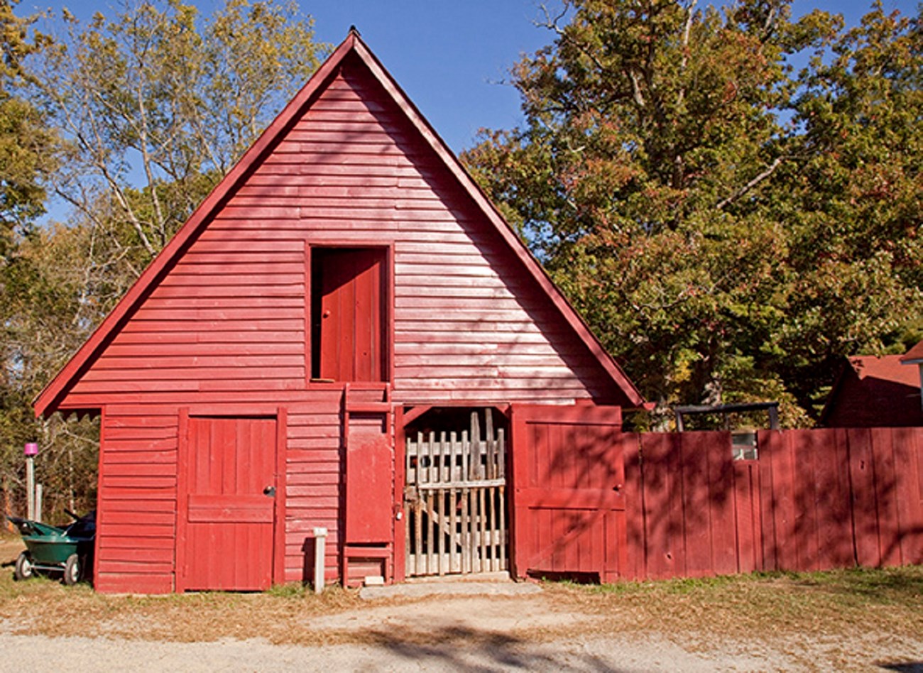A small red A frame barn with a wooden fenced play yard