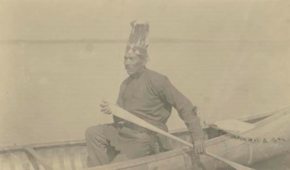 lt= An elderly man with feathered head dress paddles a canoe in open water.