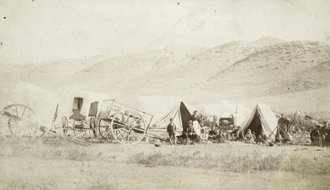 Take 2 1890_Montana HIstorical Society_Metis_p0000007 copy.tif Black and white photograph of several tents and carts. A group of people stand in front of the small camp and look towards the camera; behind them and surrounding them are tall grasses.