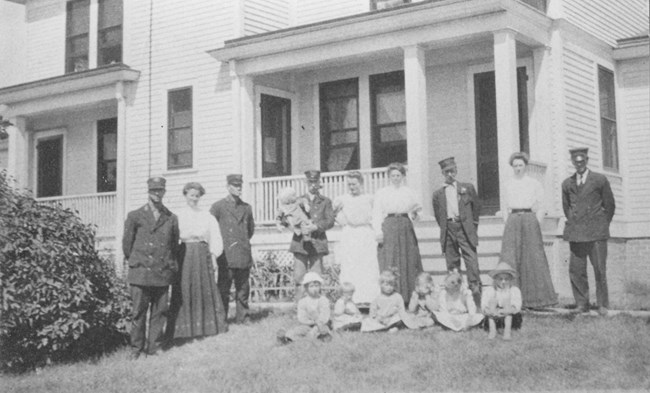 A black and white photo of 5 men in dark uniforms, 4 women in white blouses, and 7 children in the 1920's posing in front of a white house.
