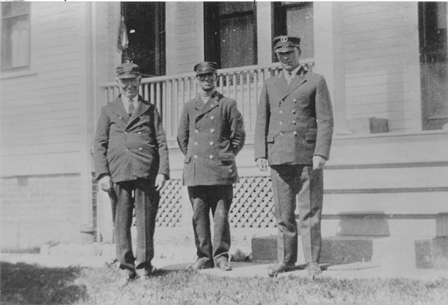 A black and white photo of three men standing in front of a house wearing a dark uniform with hats.
