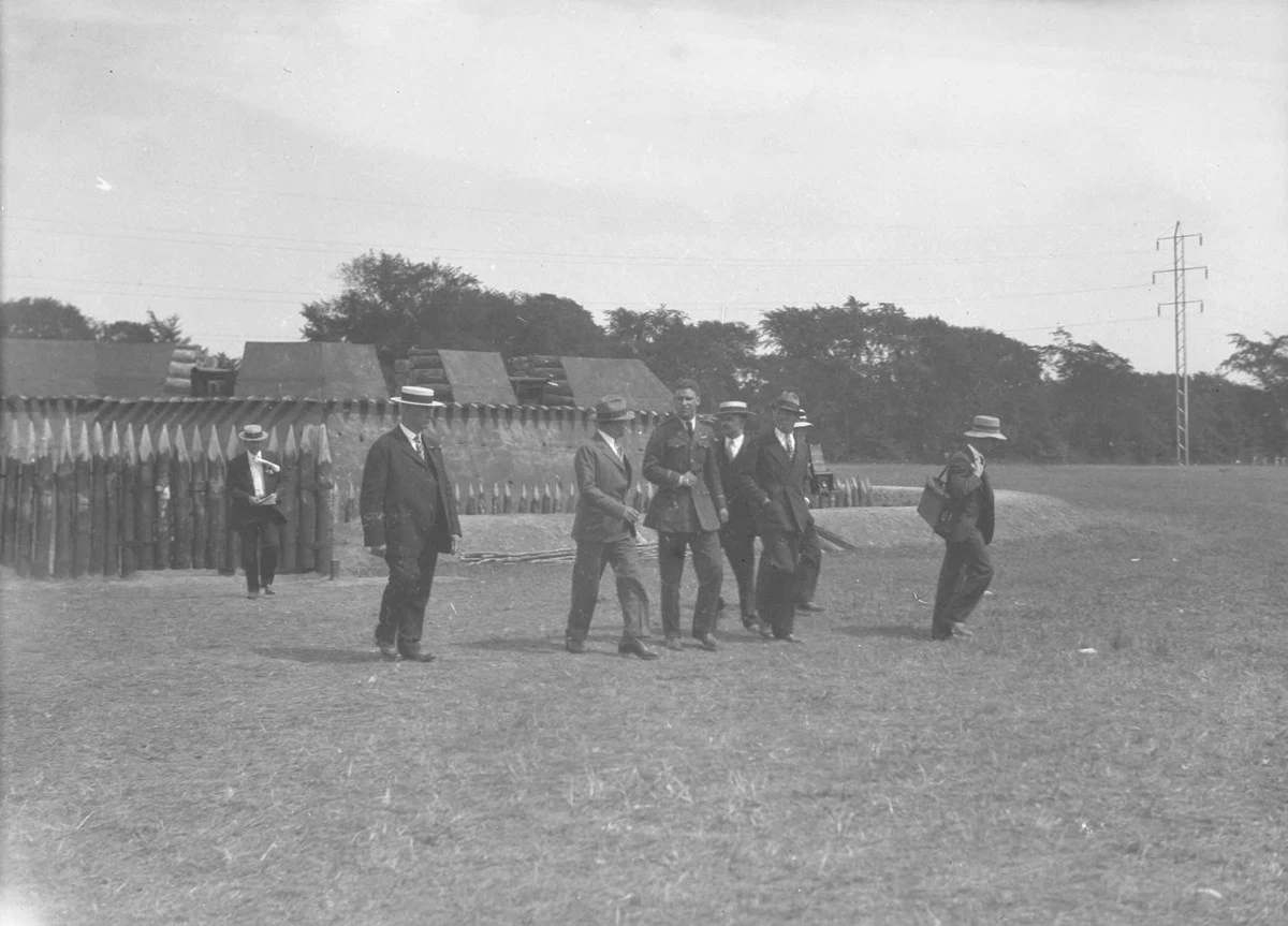 1927_Fort_1 Seven men in black suits, ties, and hats walk in front of a small wooden fort while talking to each other.