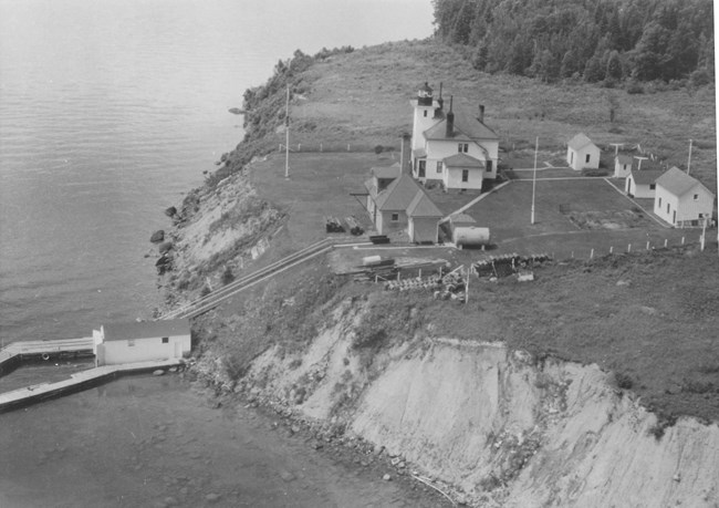 An aerial view of a white lighthouse on the steep shore of a lake, barn, fog signal building, lawn, and stairs leading down to a dock in the lake.