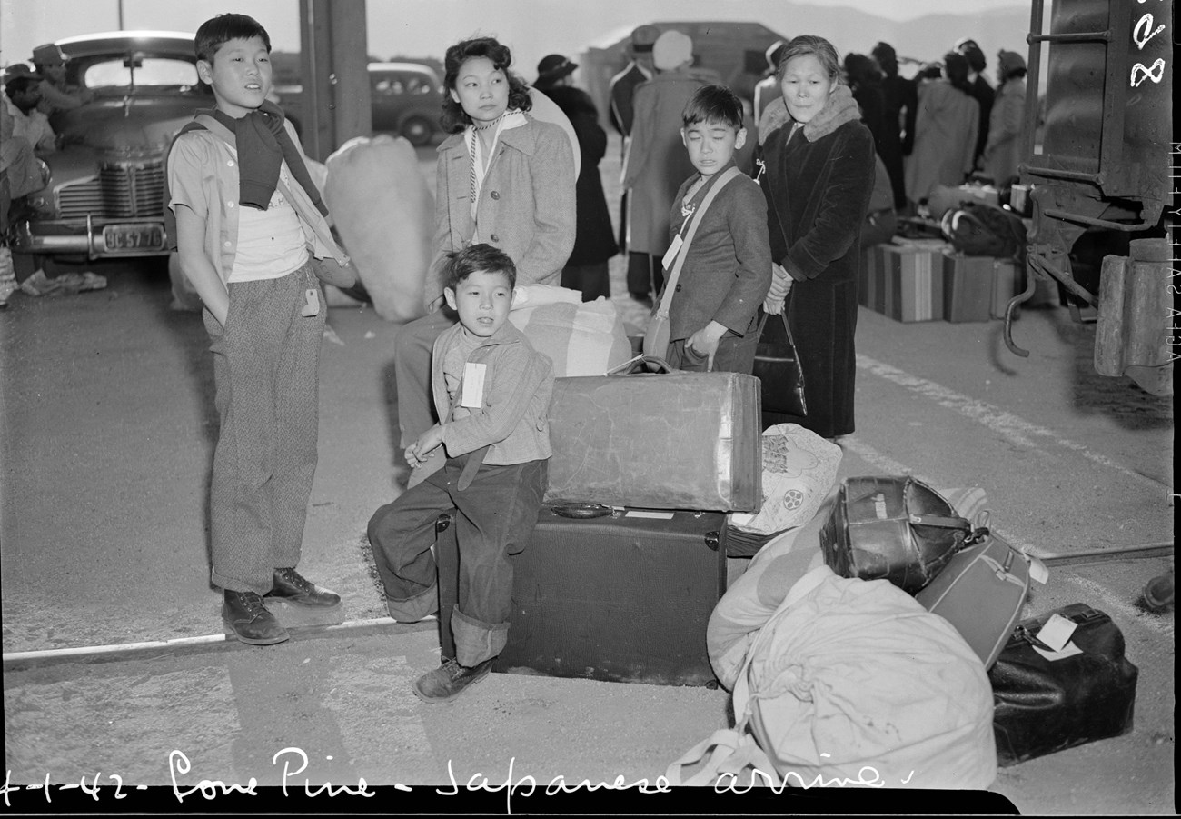 Japanese American mother, grandmother, and boys waiting with suitcases and duffel bags at train station