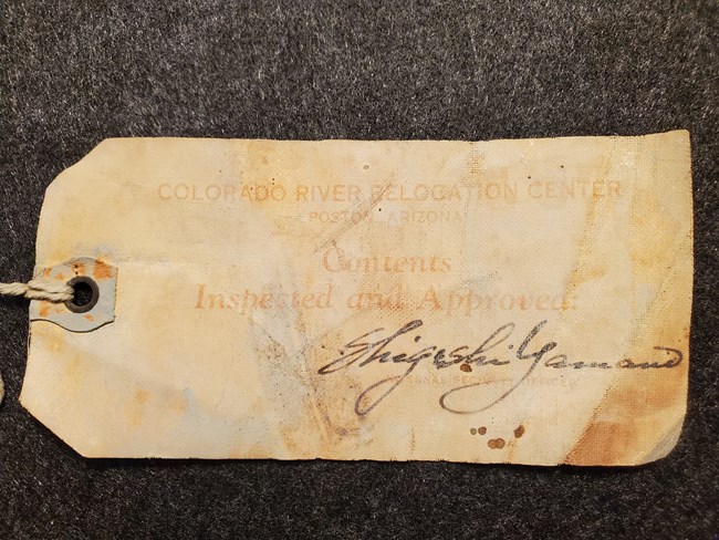 Yellowed paper luggage tag with faded text that reads “Colorado River Relocation Center, Poston, Arizona. Contents Inspected and Approved.” Shigeshi Yamano’s signature is written below the text.