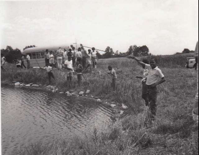 Photograph showing Summer in the Parks participants fishing. All of the participants shown in the photo are African American, and there is a young boy posing and smiling in the foreground while holding a fishing pole. There is a school bus in the backgrou