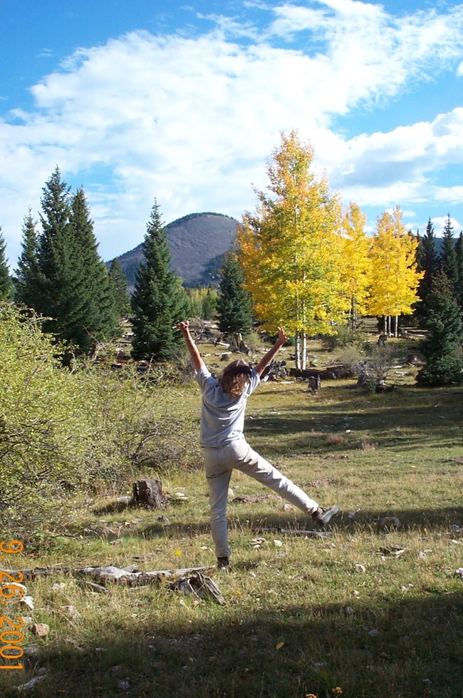 A woman with her back turned to us poses excitedly in a landscape of golden aspens and distant mountains.