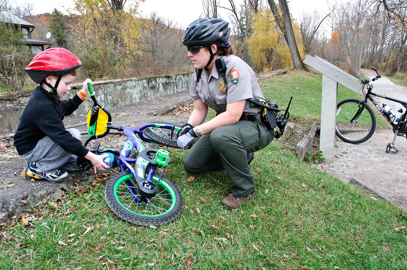 A uniformed female park ranger in black bike helmet crouches next to a young red-haired boy who holds one handlebar of the bicycle lying in the grass between them.