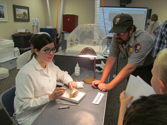 a woman in a white lab coat and a man in a park ranger uniform talk to visitors about a fossil on the table.