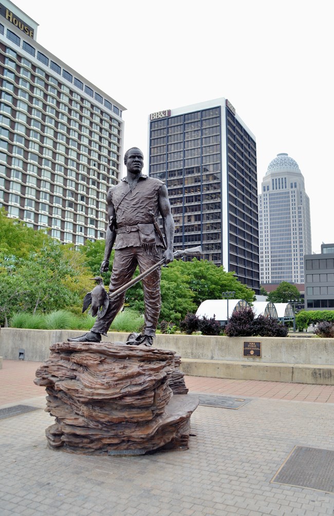 Statue of York in a plaza, city buildings visible beyond. York has a long gun in one hand and a duck in the other.
