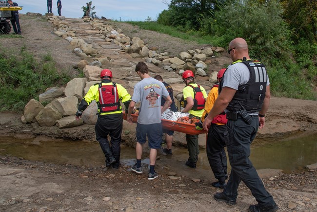 Six people in different uniforms carry an orange litter toward a stone ramp up out from a river; three of the six wear red life vests and helmets.