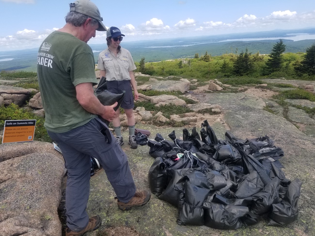 Two park volunteers in outdoor clothing carry black plastic sandbags on a granite summit,