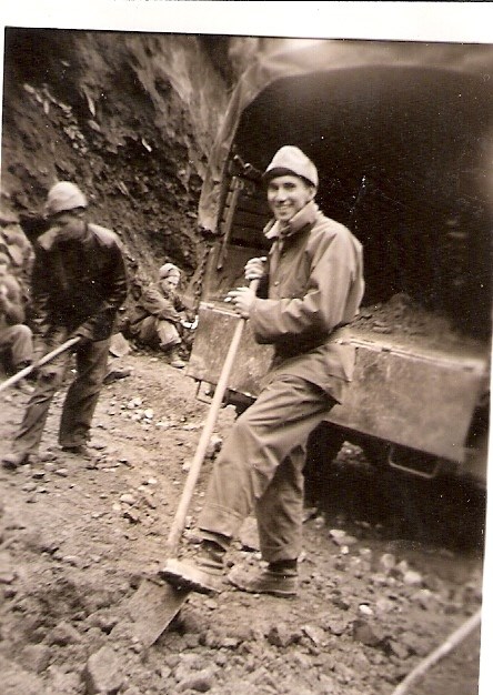 Two men with shovels work on a road, with a third, seated man, in background, and a truck.