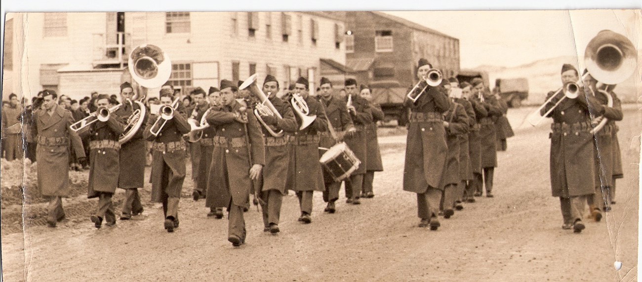 Group of men in uniform playing brass instruments marching towards the camera, down a street.