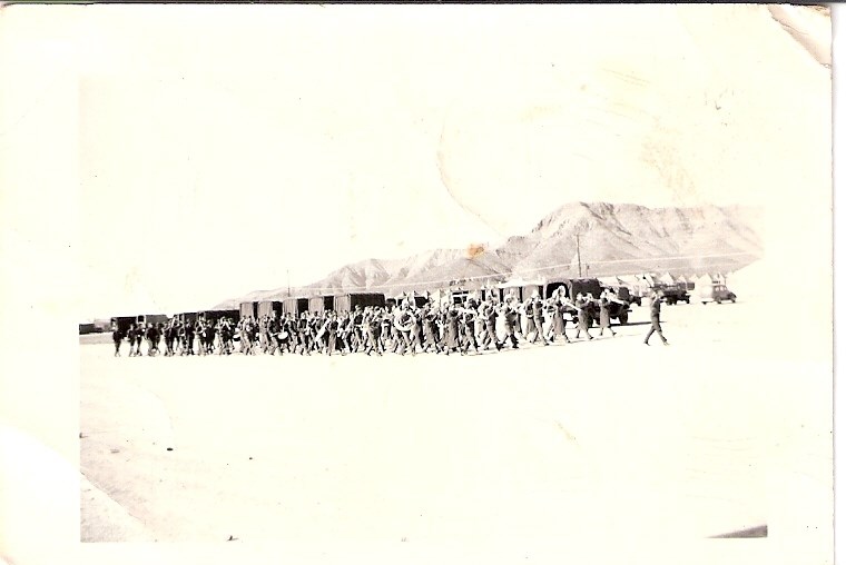 large group of men with musical instruments on parade in desert landscape with mountains and tents in background