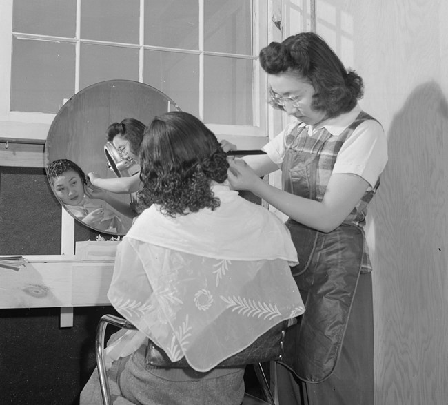 A woman standing trims the hair of a seated Japanese American woman in a room with bare wooden walls